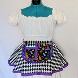 A white ruffled blouse with puffed short sleeves and a black-and-white harlequin skirt, styled on a teal mannequin torso. A colorful embroidered apron-style pocket with purple trim is attached at the waist.